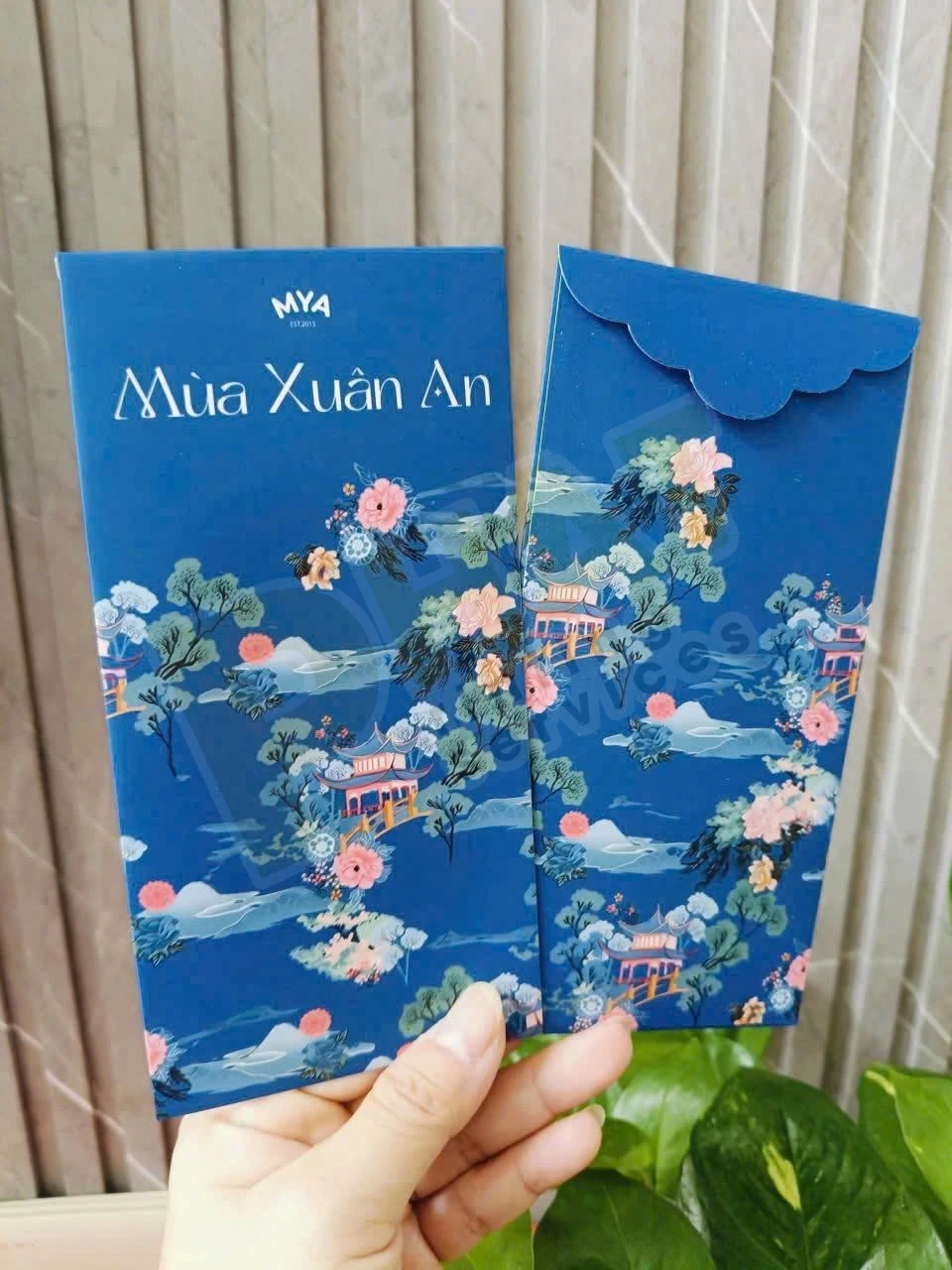 Tay cầm hai bao lì xì nền xanh đậm in họa tiết hoa và cảnh mùa xuân, mặt trước và mặt sau