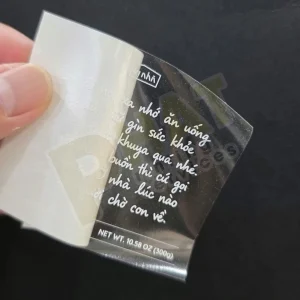 Tay cầm miếng decal trong in chữ trắng nét viết tay, đang bóc khỏi lớp đế trên nền đen