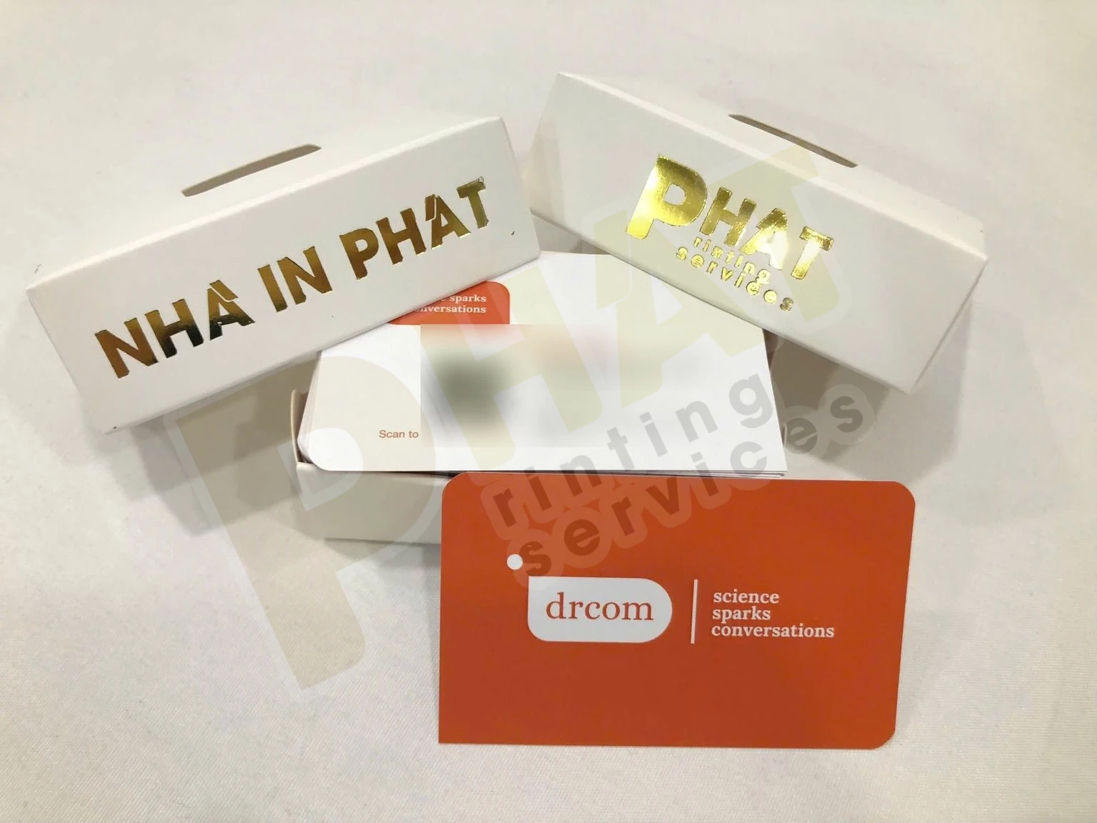 Xấp name card trắng trong hộp giấy có chữ ép nhũ vàng, phía trước là một tấm name card nền cam bo góc