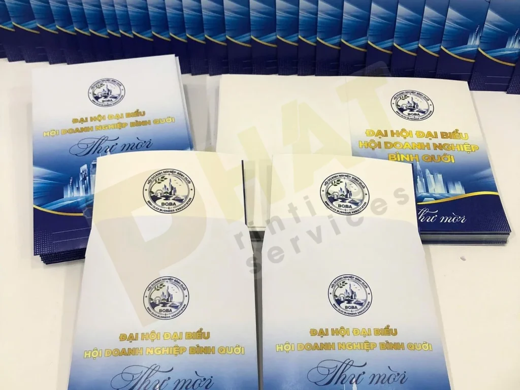 Nhiều phong bì trắng in logo và thư mời bìa xanh xếp thành hàng trên bàn