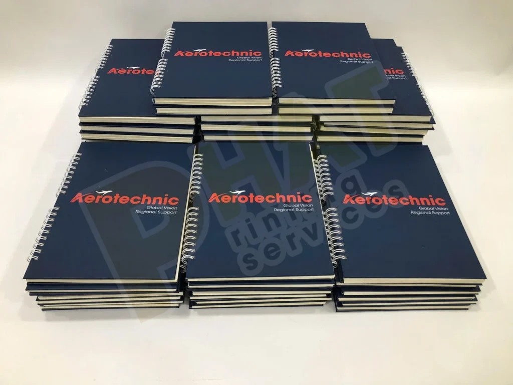 Nhiều xấp sổ tay lò xo bìa xanh đậm in logo đỏ trắng xếp trên bàn