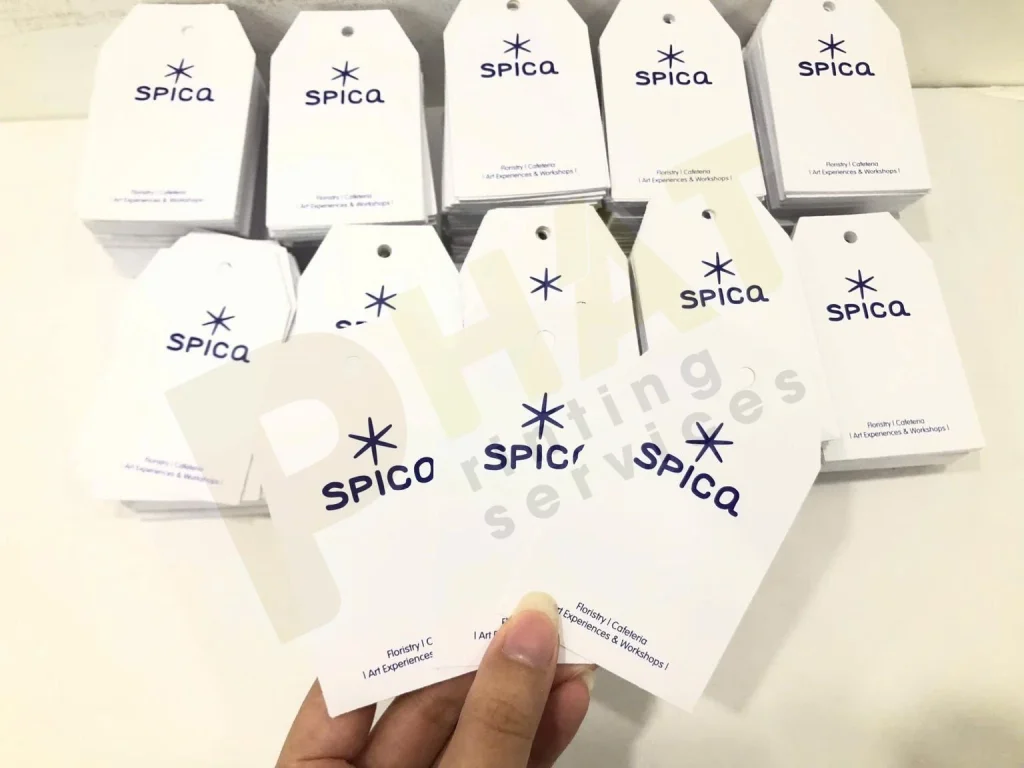In Tag Treo Quần Áo, Đồ Handmade: Kích Thước, Lỗ Xỏ Dây Và Giấy 3 Tay cầm ba chiếc tag giấy trắng có lỗ xỏ dây, phía sau là các xấp tag cùng mẫu
