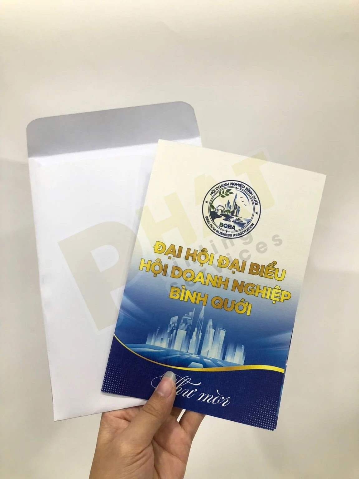 Tay cầm tấm thư mời bìa xanh in chữ vàng đặt trước phong bì trắng mở nắp