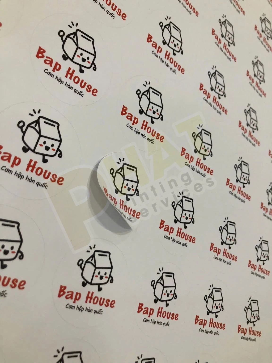 Tờ tem nhãn tròn in logo đỏ đen, một con tem được nhấc lên khỏi lớp đế