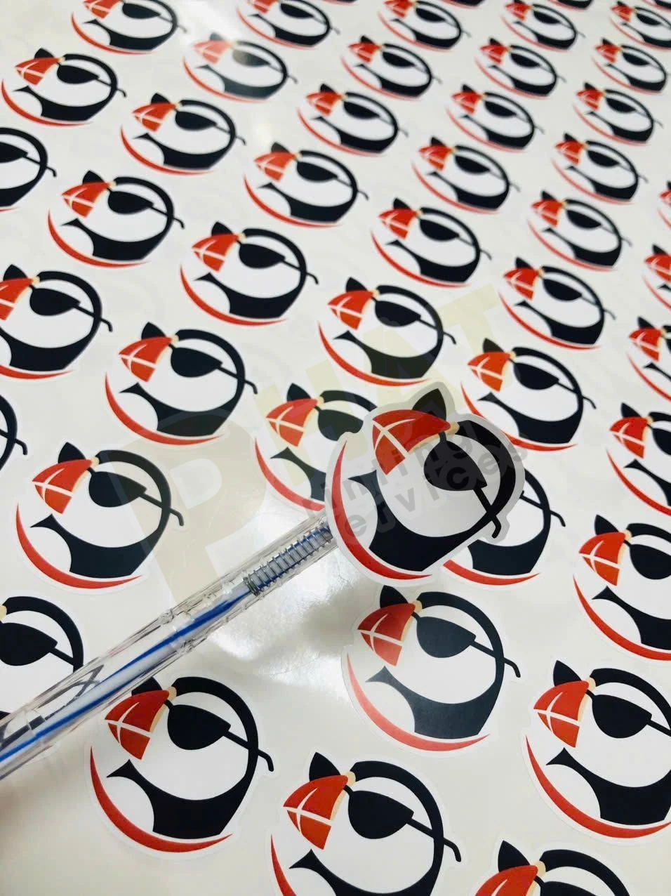 Tờ decal có nhiều con tem tròn in logo chim cánh cụt đỏ đen, một con được nhấc lên bằng bút